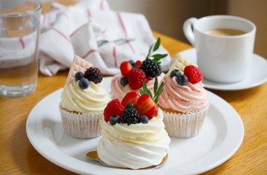 Büyük beyaz bir tabakta taze böğürtlen ve beyaz çikolatayla süslenmiş mini pavlova kekleri, bir fincan kahve, bir bardak su ve arka planda bez peçete. Seçici odak.