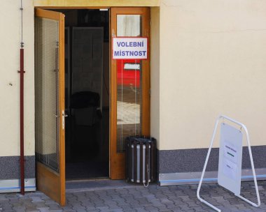 A paper sign with the Czech inscription (Eng: Polling room) stuck on the open entrance door to the building on election day in the Czech Republic, boards with voting ballots inside the polling room clipart