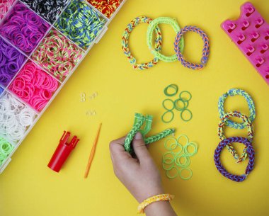 Child's hand holding a work in progress loom bracelet, other colorful elastic bracelets and bracelet making supplies on yellow background. Toy developing children's imagination. Flat lay, from above. clipart