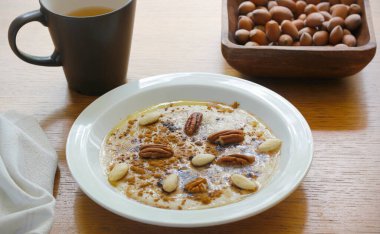 Karabuğday ve mısır irmiğinden pişirilmiş sağlıklı glütensiz yulaf lapası ve beyaz tabakta tarçın, akçaağaç şurubu, tereyağı ve fındık ile servis ediliyor. Sabah lapası, bir kupa tişört ve ahşap masada fındık..