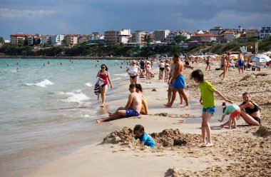Bulgaristan 'ın Primorsko kenti - 08: 29: 2024: Bulgaristan' ın Primorsko kentinde Karadeniz kıyısında dinlenen ve yüzen insanlarla dolu Sandy Southern plajı, şemsiyeler, dalgalar ve arka planda kıyı binalarıyla.