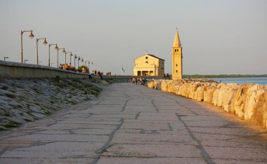 Caorle, İtalya - 08: 13: 2025: İtalya 'nın Caorle kentinde kıyı yürüyüşü, gün batımında kilise ve çan kulesiyle Madonna dellAngelo Sığınağı' na gidiyor.