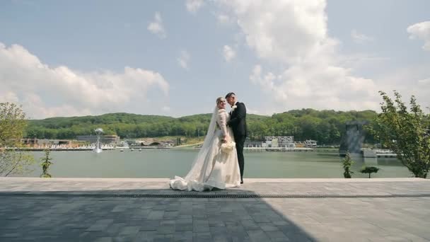キスをして 湖の近くで愛する花嫁を抱きしめる ラグジュアリーリゾートコンプレックスで結婚式を祝う白いドレスと黒いスーツでハッピーニューリード 愛と家族の概念 — ストック動画