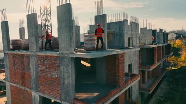 モノリスとブリックの複数の家屋の建設 ベストとハードハットの複数の労働者が建設現場の建材を梱包 — ストック動画