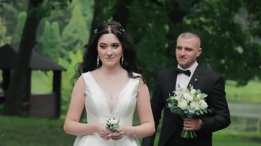 Damat, parkta bir düğün buketiyle geline yaklaşır. Gelin elinde bir yaka çiçeği tutuyor. Gelin ve damadın parkta nikah yürüyüşü.