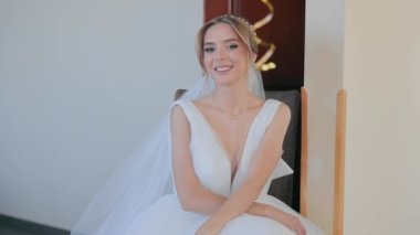 Güzel bir gelin oturuyor ve kameraya bakıyor, içtenlikle gülümsüyor. Düğün günü için hazırlık yapıyorum. Mutlu gelin.