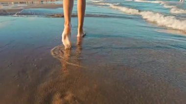 Zayıf kızların ayakları ve bacakları kumlu sahilde yürüyor. Orada su damlaları, dalgaların köpüğü kızların bacaklarını yıkıyor. Kumsalda kumsalda yürür