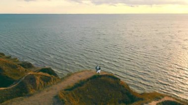 Denize yakın bir tepede duran genç bir çift, resimli bir resim. Beyaz elbiseli bir kız kocasının elini tutarak yürür, rüzgar elbiseyi farklı yönlere savurur. Hava görünümü