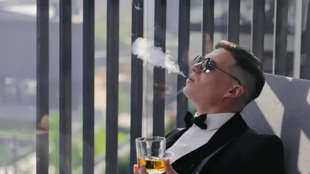 一位穿着黑色西服 戴着黑色眼镜的优雅男子坐在阳台上 手里拿着一杯酒精 抽着烟 呼着烟 新郎为艰难的婚礼做好准备 — 图库视频影像