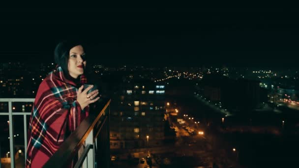 一个微笑的女人站在阳台上 裹着温暖的毯子 端着一杯热咖啡或茶 在灯光下看着美丽的夜城 一位中年妇女晚上站在阳台上 — 图库视频影像