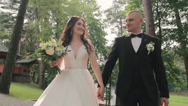新婚夫婦は結婚式の日に公園を歩いている 美しい花嫁と花嫁は 愛し合って手を握り 笑顔で互いを見つめ合っています 公園内の新婚旅行のウェディングウォーク — ストック動画