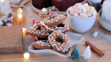 Zencefilli kurabiyeler beyaz tabakta yatar. Kurabiyeler ve bir fincan sıcak çikolata. Etrafı Noel ışıklarıyla çevrili ahşap bir masanın üzerinde marşmelov.