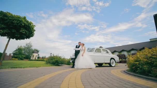 新婚旅行者はレトロカーの近くに立って 青空の背景にキスします 新婚旅行者の結婚式の日 カメラが彼らの上を走り 彼らはカメラのためにポーズします — ストック動画