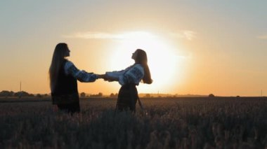 Gün batımında iki kız buğday tarlasında dans ediyor. Ulusal giysili iki kız altın buğday tarlasında el ele dans ediyor.