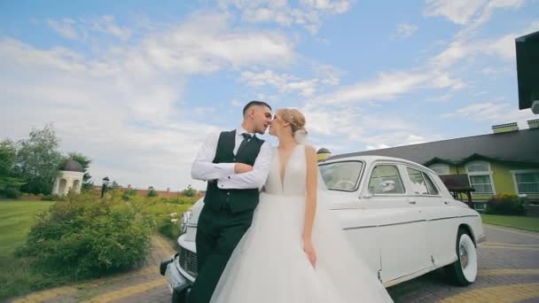 新婚旅行者はレトロカーの近くに立って 青空の背景にキスします 新婚旅行者の結婚式の日 カメラが彼らの上を走り 彼らはカメラのためにポーズします — ストック動画