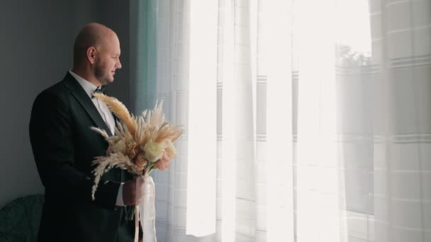 グロムホールディングウェディングブーケ 素朴な結婚式の花束を保持する黒いスーツで思慮深いグロム 明るい窓を眺める — ストック動画