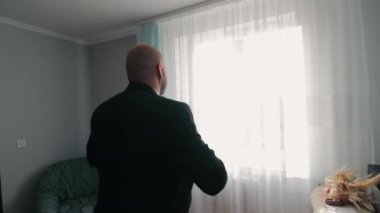 Dalgın adam perdelerden dışarı bakıyor koyu renk gömlekli kel bir adam perdelerle örtülü parlak bir pencereden dışarı bakıyor.