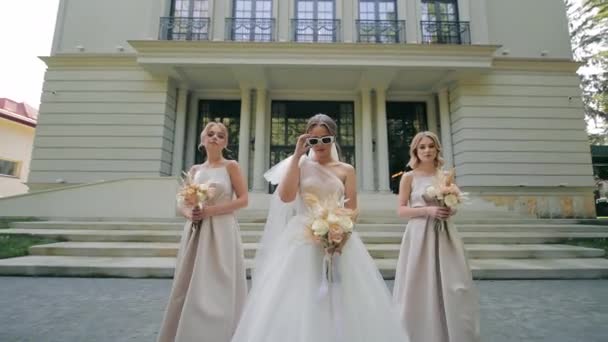ブライダルエレガンス 花嫁の花嫁と花嫁の花嫁 白いガウンとサングラスのスタイリッシュな花嫁は グランドビルの外にベージュドレスで2つの花嫁の間に立っています — ストック動画