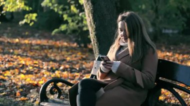 Şehir parkında sonbahar sükuneti, güneşli bir sonbahar gününün tadını çıkaran genç bir kadın, bir bankta oturuyor ve akıllı telefonunu kullanıyor..