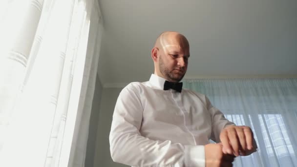 绅士调整手表事件发生前 一位穿着白衬衫的体贴男子调整了领结 沐浴在柔和的阳光下 沐浴在窗边 — 图库视频影像