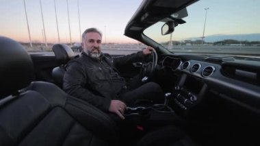 Bir adam gün batımında üstü açık, rahat bir ifadeyle üstü açık bir araba süren lüks, olgun bir adamın içinde oturur.