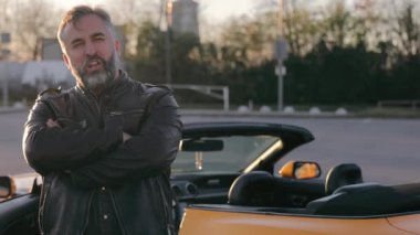 Üstü açık bir arabanın yanında deri ceketli bir adam. Portre, dayanıklı bir adam gün batımında üstü açık bir arabanın yanında kollarını kavuşturur..