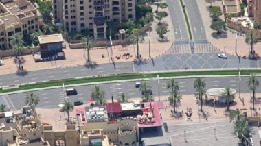 Dubai 'deki işlek kavşak ve sokak pazarının hava görüntüsü, modern yollar ve verimli palmiye ağaçları ile birlikte develerin ve market tezgahlarının bulunduğu hareketli bir sokak sahnesinin yüksek açılı çekimi.