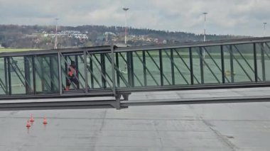 Havaalanı Terminali 'nde çalışan işçiler, yüksek görüş mesafesi olan turuncu üniformalı işçiler havaalanının cam yolu boyunca yürüyor, yoğun bir havaalanını ve arka planda yemyeşil bir manzarayı yansıtıyor.