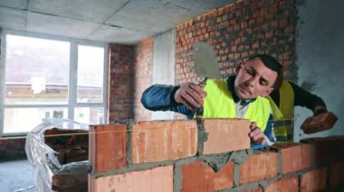 Tuğla Duvar inşa eden inşaat işçisi, yansıtıcı yelek içindeki odaklanmış bir inşaat işçisi titizlikle yenileme aşamasındaki bir binanın içinde kısmen inşa edilmiş bir duvara tuğla örüyor.