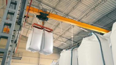 Endüstriyel vinç geniş bir depoda büyük beyaz çantaları kaldırıyor. Sahne vinçlerin mekanik yapısını ve deponun yüksek tavanını yakalıyor..