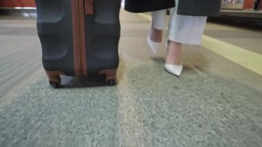 Bir gezgin, havaalanı terminalinde yürürken siyah bir bavul çeker. Görüntü, kişinin alt yarısını yakalar ve şık beyaz ayakkabılarını ve kıyafetinin bir kısmını gösterir..