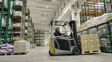 Bir depo işçisi büyük, organize bir tesiste forklift kullanıyor. Olay yerinde çeşitli ürün ve malzemelerle dolu raflar bulunuyor..