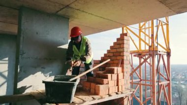 İnşaat işçisi Tuğla Duvar, kırmızı şapka ve sarı güvenlik yeleği giyen bir inşaat işçisi inşaat sahasına tuğla duvar örüyor..