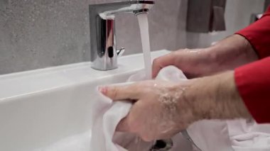 Lavaboda akan suyla beyaz giysileri yıkayan ellerin yakın çekimi. Erkekler lavaboda akan suyun altında beyaz bir gömlek yıkar.