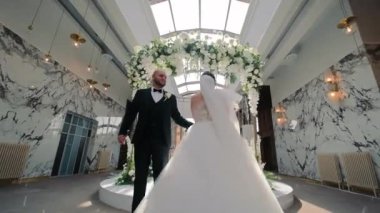 Gelin ve damat bir çiçek kemerinin altında dans ediyorlar. Damat siyah bir smokin ve duvak takmış beyaz bir gelinlik giyiyor. Arkaplanda mermer duvarlar ve modern ışıklandırma yer alıyor.