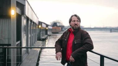 Sakallı bir adam nehir kenarında duruyor, korkuluklara yaslanıyor, modern binalar ve alacakaranlıkta nehir var.