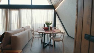Modern ahşap bir kulübede, parlak ve davetkar bir yemek salonu. Yuvarlak bir masa, şık sandalyeler, bir çiçek süsü ve geniş pencerelerden manzaralı bir manzara.