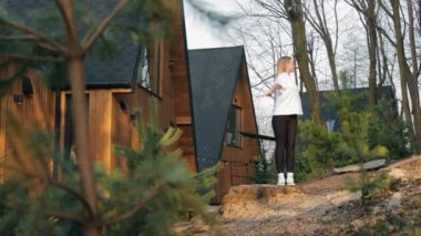 Ağaçlarla çevrili ahşap bir kulübede sabah egzersizi yapan bir kadın, temiz havanın ve sabah ışığının tadını çıkarıyor, doğal ortamdaki sağlıklı ve aktif yaşam tarzını simgeliyor..