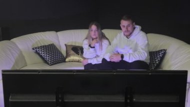 Çift beyaz bir kanepede birlikte televizyon izliyor, çift beyaz bir kanepede yan yana oturuyor, loş bir odada dikkatle televizyon seyrediyor..