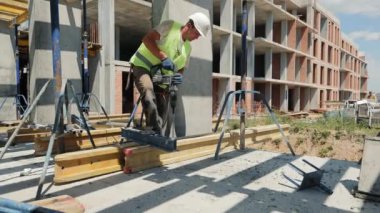 İnşaat işçisi inşaat sahasında sondaj yapıyor. Bir inşaat işçisi inşaat alanında yüksek görüş mesafesi olan bir yelek ve sıkı bir şapka takarak beton bir levhayı delerken görülüyor.