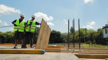 İnşaat işçileri inşaat alanında büyük bir kurul kuruyor. Yüksek görüşlü yelek ve şapka giyen iki inşaat işçisi bir inşaat sahasına büyük bir ahşap tahta yerleştirmek için birlikte çalışıyorlar.