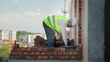 Mason inşaat alanında tuğla duvar üzerinde çalışıyor. Yüksek görüşlü yelek giyen bir inşaat işçisi inşaat sahasının üst katlarına tuğla duvar örmeye odaklanır.