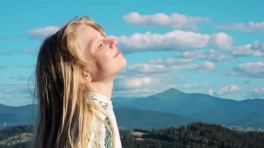 Arka planda dağlarla gün ışığının tadını çıkaran uzun sarı saçlı genç bir kız gözleri kapalı güneşin tadını çıkarıyor.,