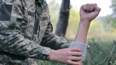 Dışarıdaki bir kola bandaj yapıştıran bir asker. Kamuflaj üniformalı bir askerin dış mekânda sargı bezi sararken çekilmiş yakın plan bir fotoğrafı.