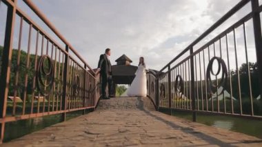 Kırsal bir köprüde, arka planda çardak olan bir çift. Yeni evli bir çift kırsal bir köprüde duruyor, doğal yeşilliklerle çevriliyken birbirlerine bakıyorlar.