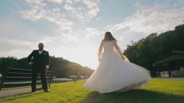 Gelin ve Damat, Sunlit Anı 'nın tadını çıkarıyor. Gelin, beyaz elbisesiyle çimenlerin üzerinde zarif bir şekilde dans ederken damat, her ikisi de altın bir öğleden sonra güneşinin altında ayakta duruyor.