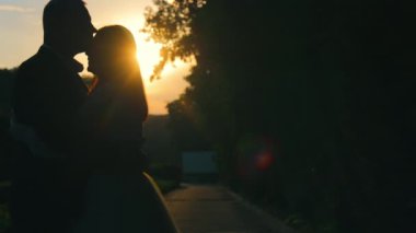 Gelin ve Damat Açık havada Günbatımı Öpücüğü Paylaşıyorlar. Bir gelin ve damat, güneş arkalarında batarken sıcak bir altın parıltı saçarak açık havada samimi bir anı paylaşıyor.