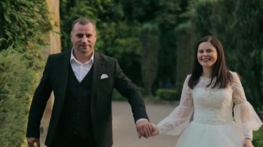 Mutlu bir düğün çifti el ele tutuşuyor ve dışarıda yürüyor beyaz dantelli bir gelinlik içinde gülen bir gelin ve siyah takım elbiseli bir damat el ele tutuşup dışarıda birlikte yürüyorlar..