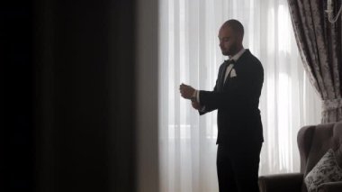 Damat pencerenin yanında duruyor kol düğmelerini ayarlıyor, parlak siyah smokinli bir damat güneş ışığında duruyor, kol düğmelerini ayarlıyor.,