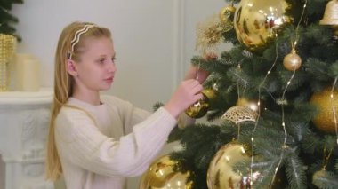 Noel ağacını altın süslemelerle süsleyen kız, krem rengi süveter giyen genç kız güzel dekore edilmiş bir Noel ağacına parlak altın süsler asıyor.